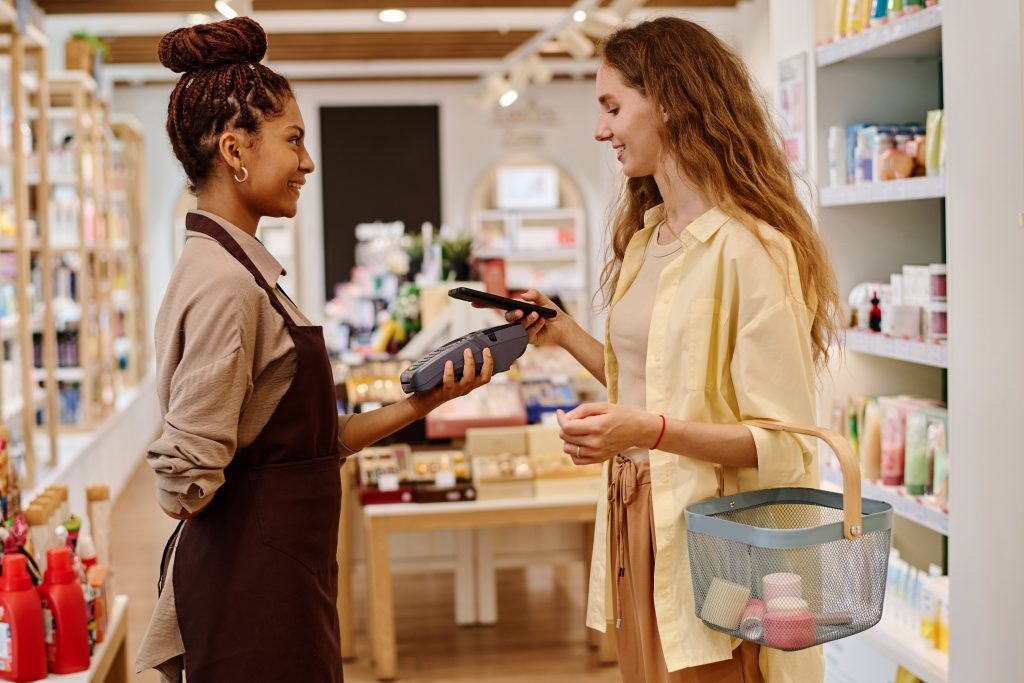 Young woman using smartphone to pay for her purchase while saleswoman holding terminal for payment