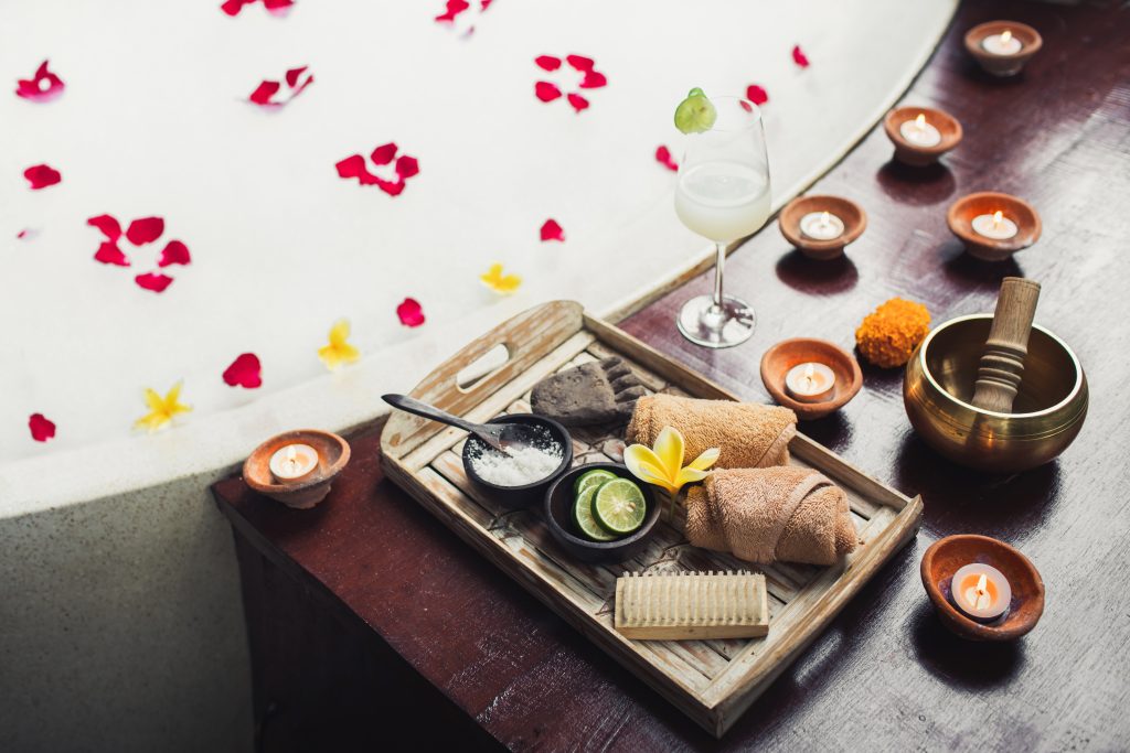 Romantic spa set with lime, scrub, salt, towels and brush in wooden tray. Flower petals in bath tub with foam and candles around. Organic and natural beauty treatment assesory.