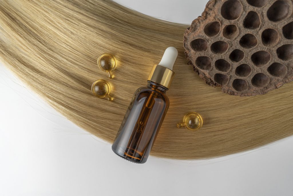 A hair care oil or serum in a brown dropper bottle and golden capsules lying on a blond hair strand, product marketing mockup