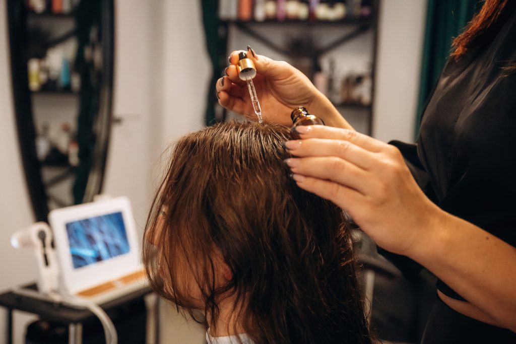 Drips serum onto head, hair roots. Woman is in a beauty salon with hair stylist.