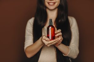 Studio portrait of happy satisfied woman holding her long healthy brown hair and showing at camera shampoo or conditioner, female using natural paraben free hair-care beauty products, selective focus