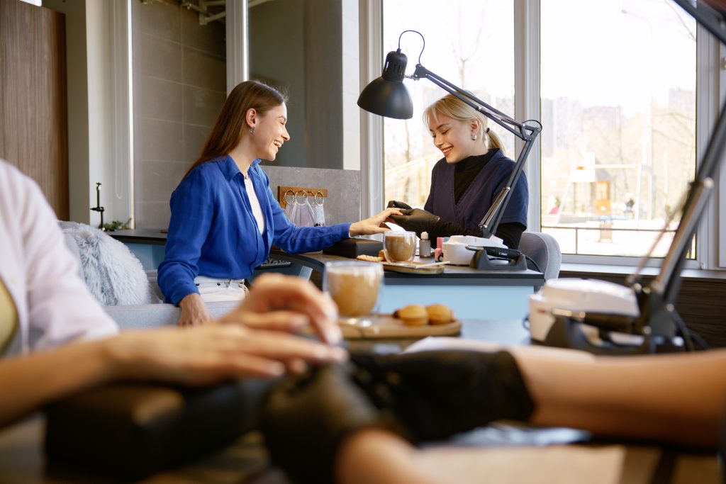 Female client getting high quality service at nail studio. Young woman customer drinking cappuccino eating sweet snacks while manicurist doing manicure
