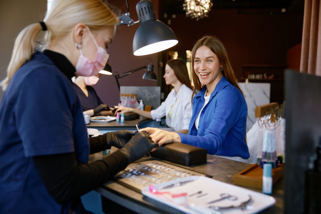 Smiling woman client enjoying manicure procedure at beauty studio. Professional manicurist in protective mask and gloves doing nail treatment for satisfied female customer