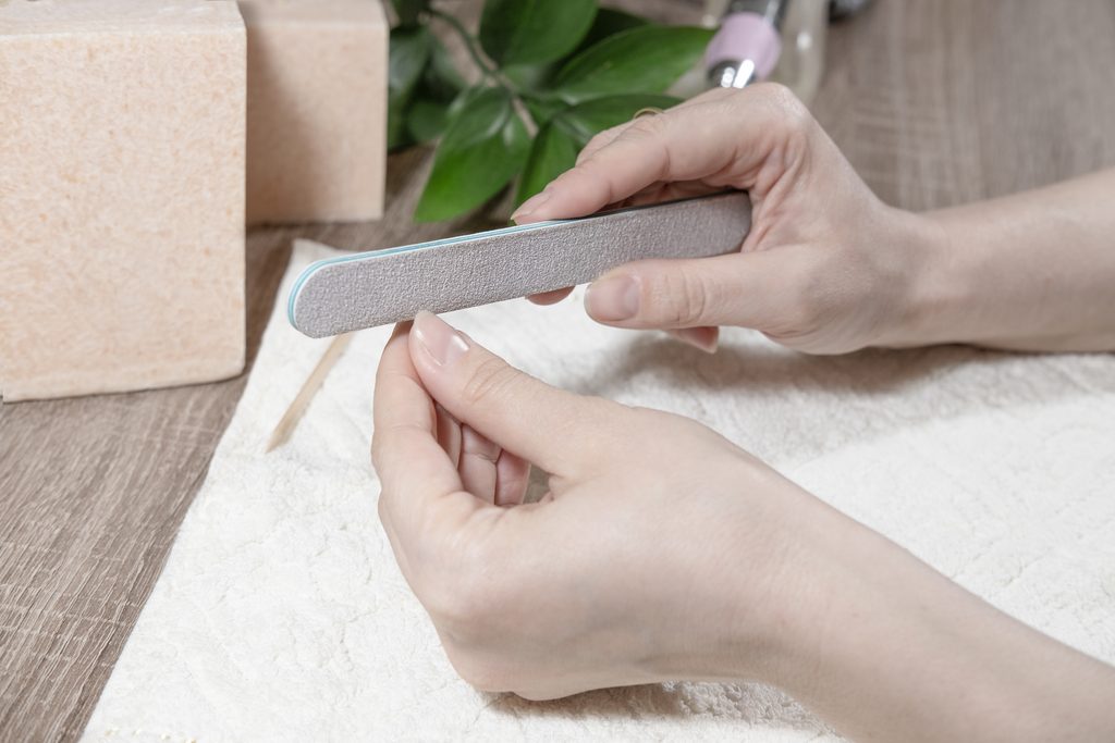 A woman manicurist treats her nails with a nail file, polishes the surface of the nail, smoothes the shape of the nail in a beauty salon. Manicure process close-up.
